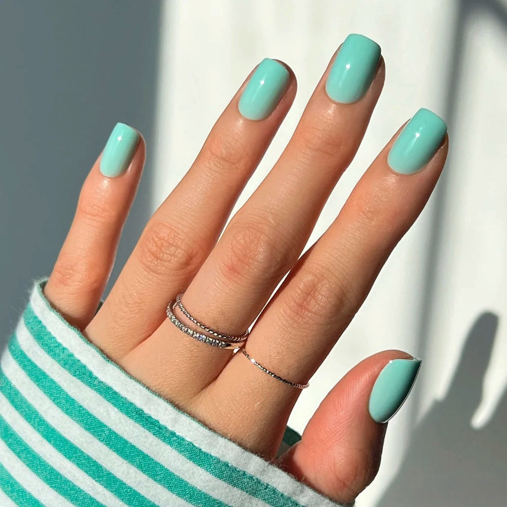 A hand with glossy Soft Teal squoval nails, wearing two thin metal rings.