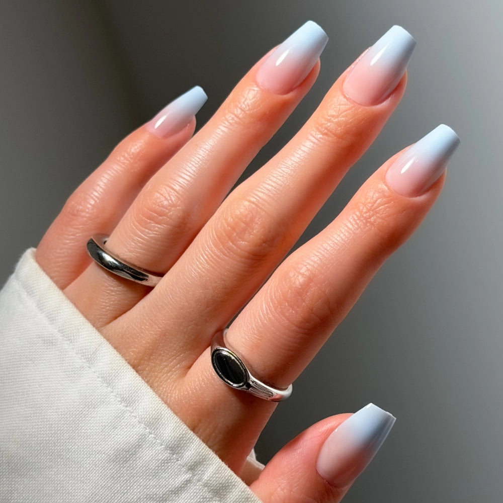A hand with long, square-shaped “Soft Skyline” nude-to-blue ombré press-on nails by glamnetic wears two silver rings and a white sleeve. - Product image