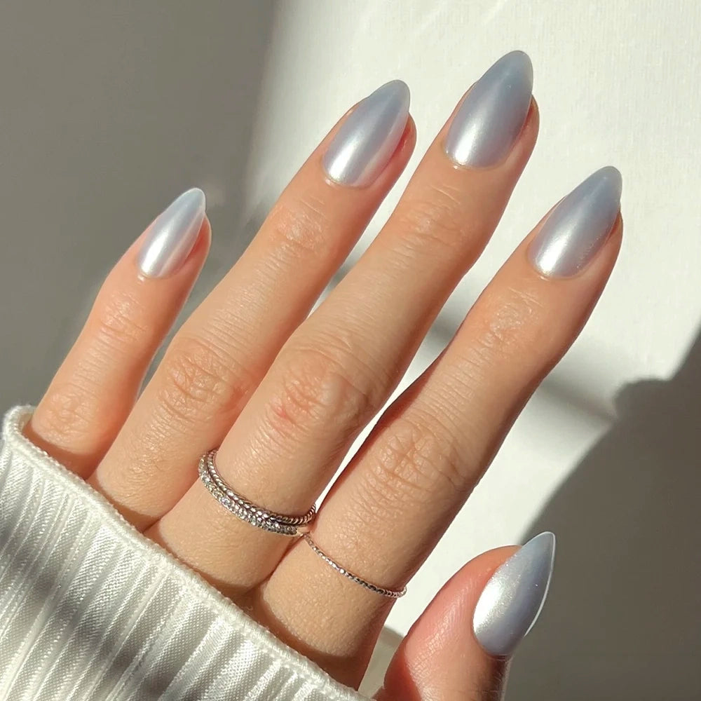 A hand wearing glamnetic Something Blue press-on nails, which are short, almond-shaped, and metallic baby blue, accessorized with two thin rings.