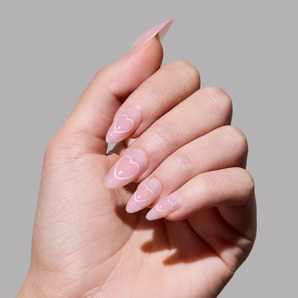 A hand with long, almond-shaped nails showcases glamnetic’s Soulmate Energy—a soft nude-pink velvet manicure with cat-eye heart designs outlined in white on a shimmery, translucent pink base, set against a plain gray background. - Product image