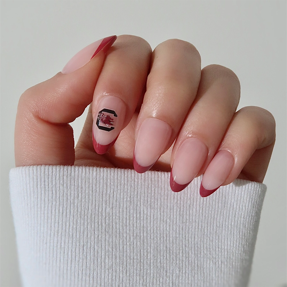A hand showcases the glamnetic South Carolina Gamecocks Press-On Nail Set, featuring almond-shaped nails with pink polish, dark red French tips, and a Gamecocks logo accent. The look is paired with a white long-sleeve shirt. - Product image