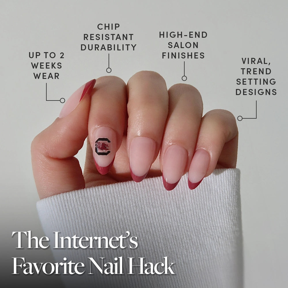 A hand with almond-shaped South Carolina Gamecocks Press-On Nails by glamnetic features pink polish, red tips, and a black accent design. Enjoy salon-quality, chip-resistant wear. A white sleeve partially covers the hand. - Product image