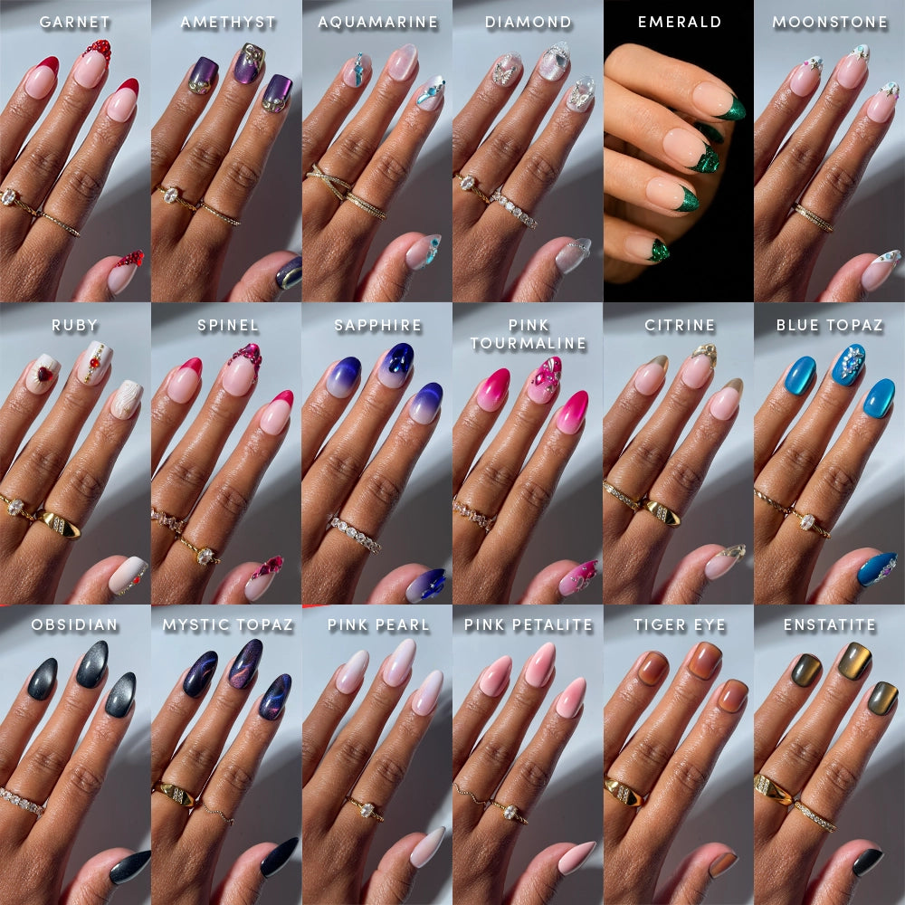 A hand showcasing the "Sparkling Gems Collection" by glamnetic, featuring intricately painted nails inspired by gemstones and labeled with names such as garnet, amethyst, aquamarine, diamond, emerald, moonstone, ruby, spinel, sapphire, pink tourmaline, citrine, blue topaz, obsidian, mystic topaz, pink pearl, pink petalite, tiger eye and enstatite.