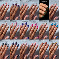A hand showcasing the "Sparkling Gems Collection" by glamnetic, featuring intricately painted nails inspired by gemstones and labeled with names such as garnet, amethyst, aquamarine, diamond, emerald, moonstone, ruby, spinel, sapphire, pink tourmaline, citrine, blue topaz, obsidian, mystic topaz, pink pearl, pink petalite, tiger eye and enstatite.