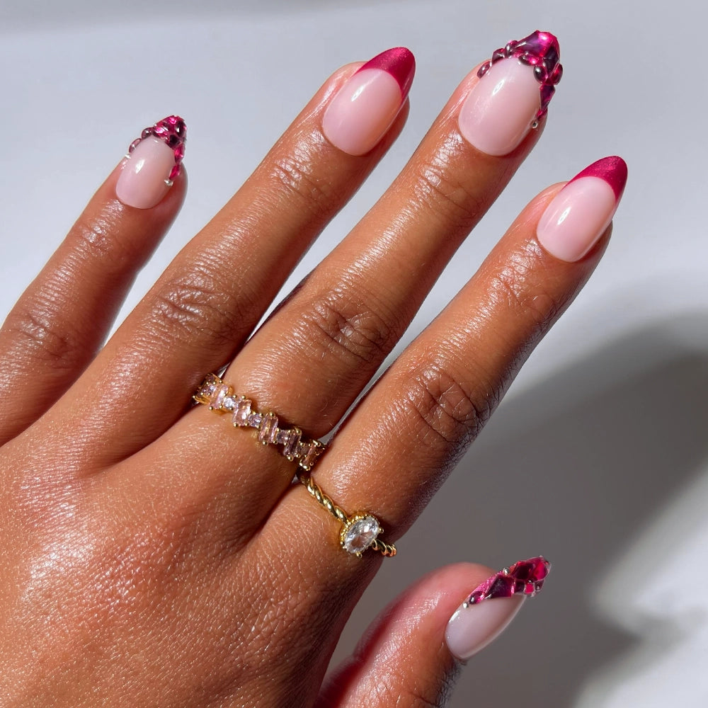 A hand showcases the "Spinel" French tip press-on nails by glamnetic, featuring almond-shaped pink nails with rhinestones along the tips for a salon-quality finish. Two gold rings adorn the fingers, highlighting the elegance of these glamorous press-on nails.