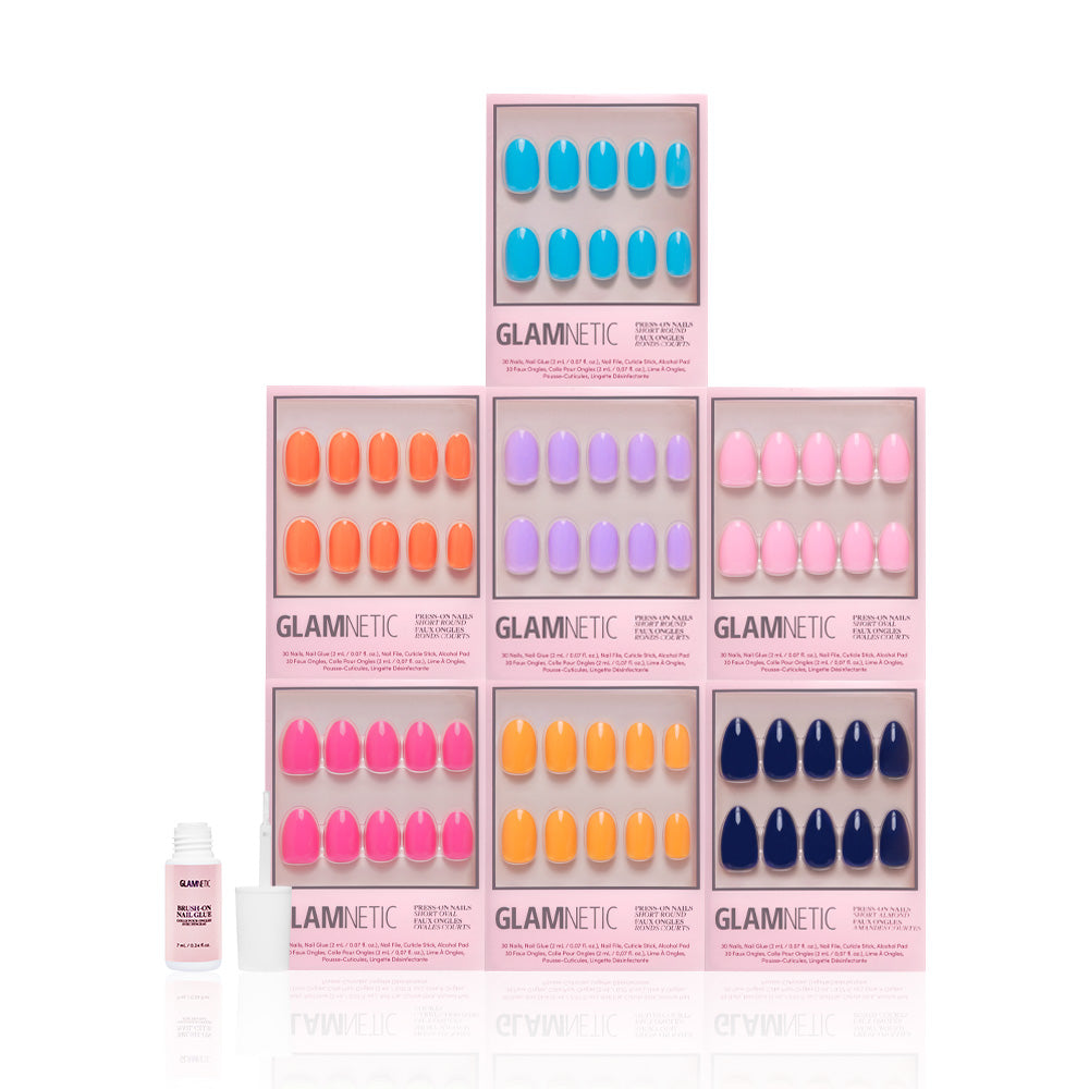 Six boxes of glamnetic Spring Sunset press-on nails, arranged in a pyramid and each showing vibrant, glossy colors. A small bottle of nail glue sits to the left, all set on a clean white background. - Product image