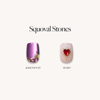 Two short squoval press-on nails from glamnetic’s Squoval Stones set are displayed: “AMETHYST” is a shimmery purple with gold and amethyst 3D art; “RUBY” is pale with a red gem and gold sunburst.