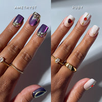 Two manicured hands with rings showcase distinct nail art: the left hand displays shimmery purple press-on nails embellished with gold and gem accents, while the right hand features glamnetic's Squoval Stones press-on nails in a short squoval shape, adorned with white polish, red gems, gold lines, and 3D heart nail art.