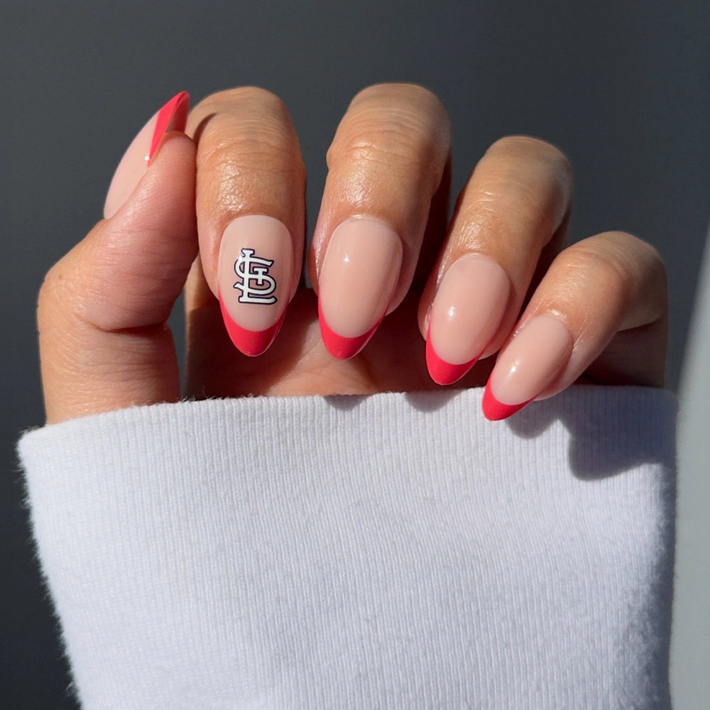 A hand features the glamnetic St. Louis Cardinals Press-On Nail Set, showcasing almond-shaped nails with a light pink base, bright pink French tips, and a white and black geometric accent. The person wears a white long-sleeve top.