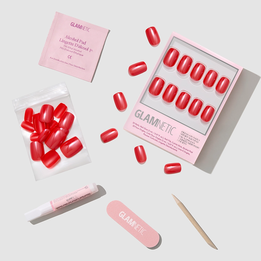 A flat lay of glamnetic's Statement Red press-on nail kit shows short squoval red nails, nail glue, a file, a wooden cuticle stick, an alcohol pad, and extra nails on a light background.