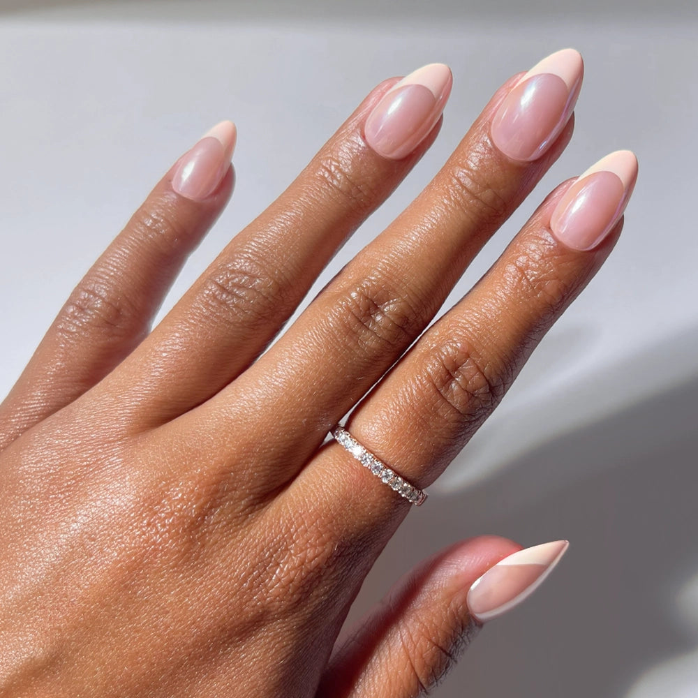 A hand with glamnetic’s Strawberry Icing short almond press-on nails in a glazed French tip style and a thin diamond ring on the ring finger, shown against a neutral background. - Product image