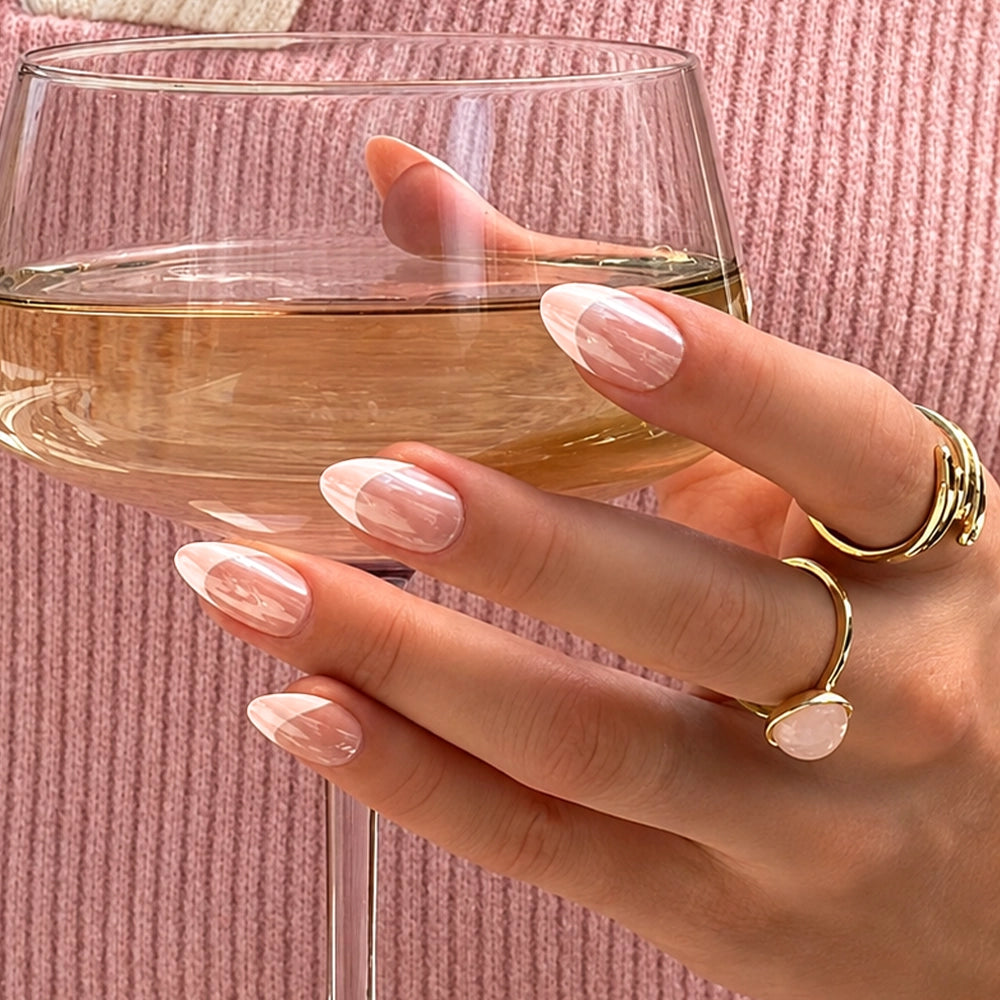 A hand with Strawberry Icing by glamnetic — pink and white glazed French tip nails shaped in a classic almond style — holds a glass of white wine. The person wears gold rings, including one with a pale pink stone, while a pink knit sweater appears in the background. - Product image