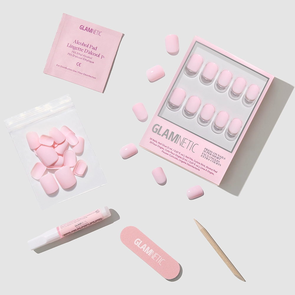 A flat lay of glamnetic’s Sugar BB kit, featuring glossy pink press-on nails, loose super short nails in a bag, an alcohol pad, nail glue, a file, and a cuticle stick—all on a light gray background. - Product image