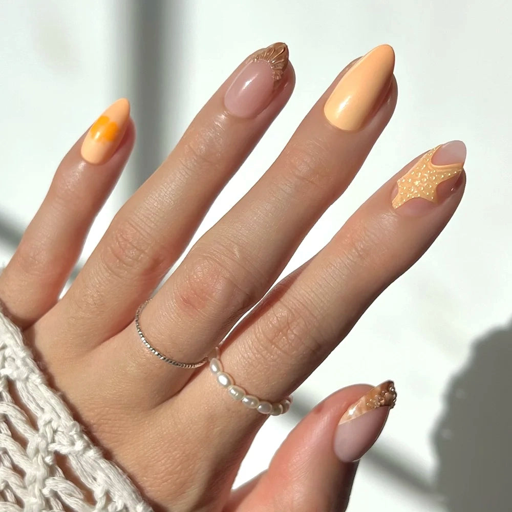 A hand with short almond nails painted in peach and nude tones features unique geometric and textured designs from the Sunny Shores set by glamnetic. Subtle hints of mermaid nail art enhance the look, while a pearl ring and a thin silver ring adorn the fingers. A white knit sleeve is partially visible.