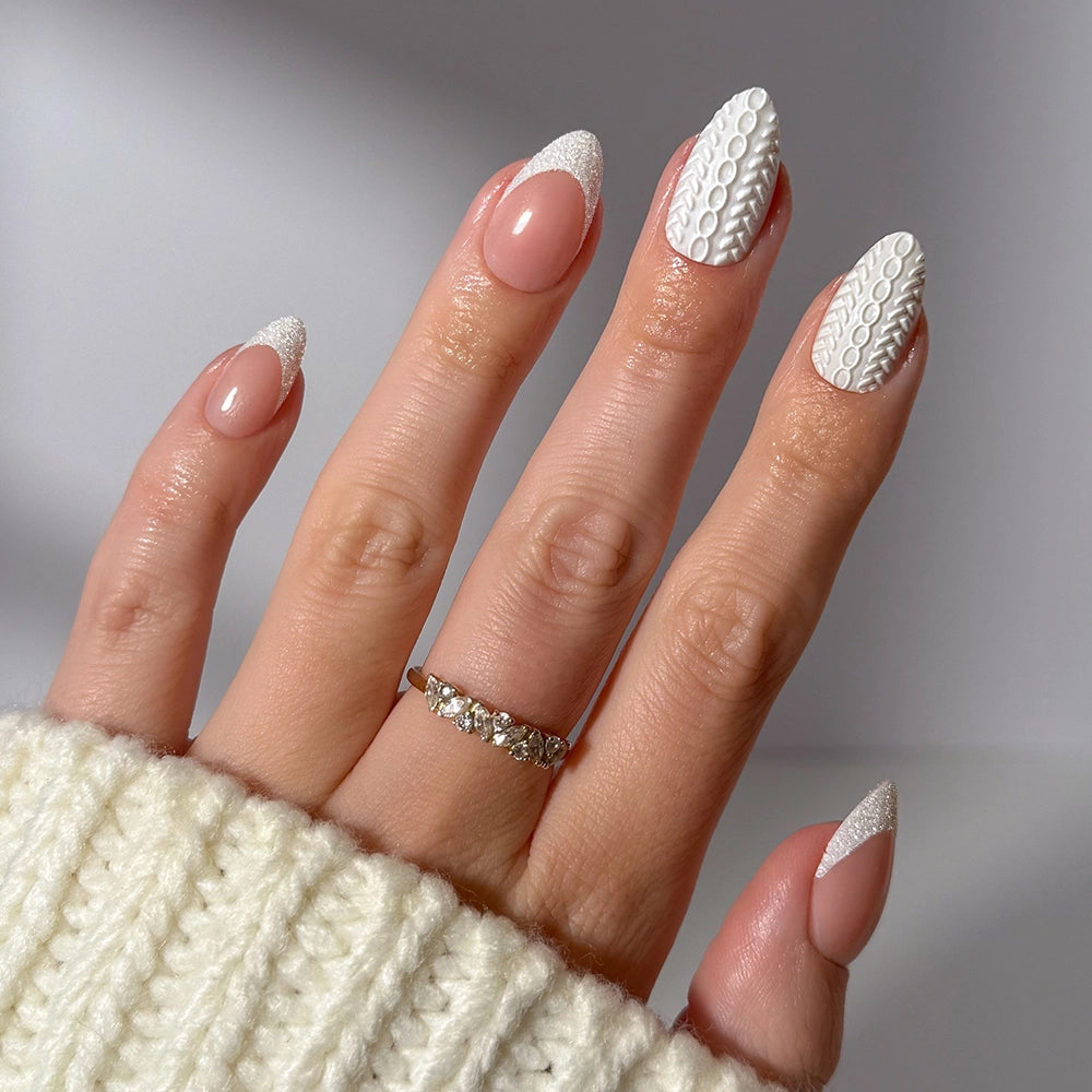 A hand with almond-shaped nails showcases glamnetic’s Sweater Weather set—nude polish adorned with white textured tips and delicate designs, offering a modern spin on classic glitter French tips. The look is paired with a diamond ring and a cream knit sweater.