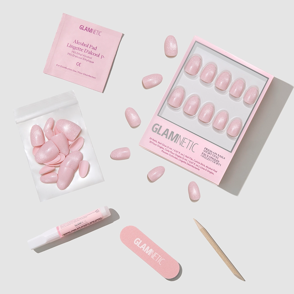 The glamnetic Sweet Talk set features short oval pink press-on nails, a nail file, cuticle stick, nail glue, alcohol pad, and extra nails—all neatly arranged on a white surface with soft shadows.
