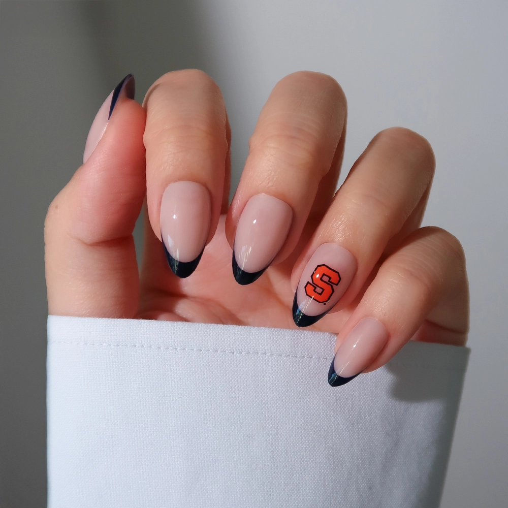 A hand with almond-shaped nails shows off the glamnetic Syracuse Orange Press-On Nail Set: nude base, black French tips, and an orange S logo on the ring finger—perfect for game day. The person is wearing a white long-sleeved shirt. - Product image