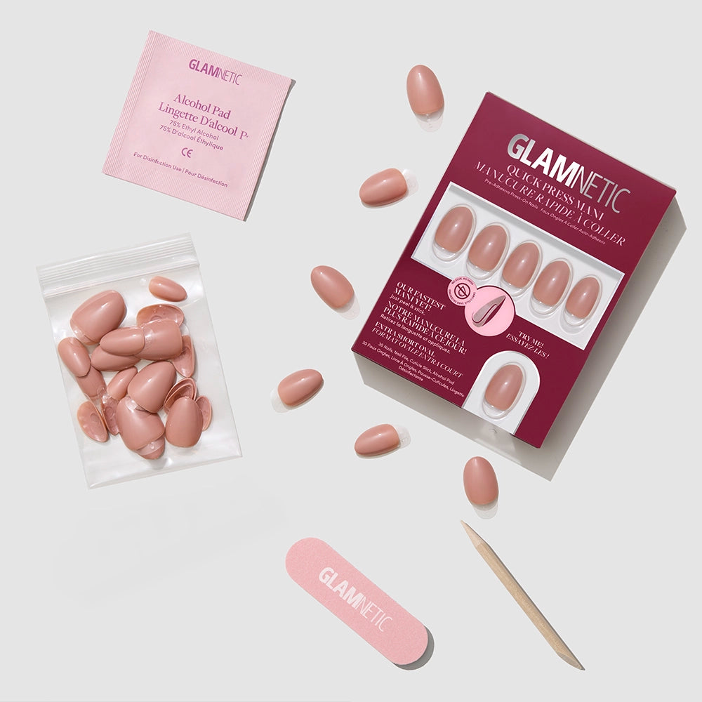 A flat lay of the glamnetic Teddy press-on nail kit featuring a box of nude pink nails, loose nails, nail glue tube, pink nail file, wooden cuticle stick, alcohol pad, and a small bag with extra nails on a light surface. - Product image