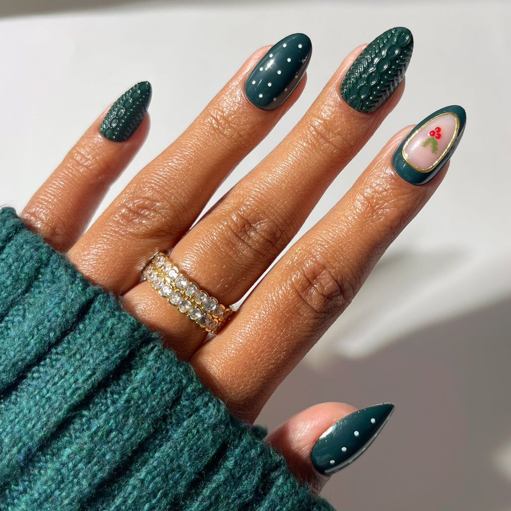 Glamnetic Table for Ten short almond press-on nails in deep green feature white polka dots, textured cable-knit patterns, and a holly berry accent on the ring finger for a festive holiday manicure.