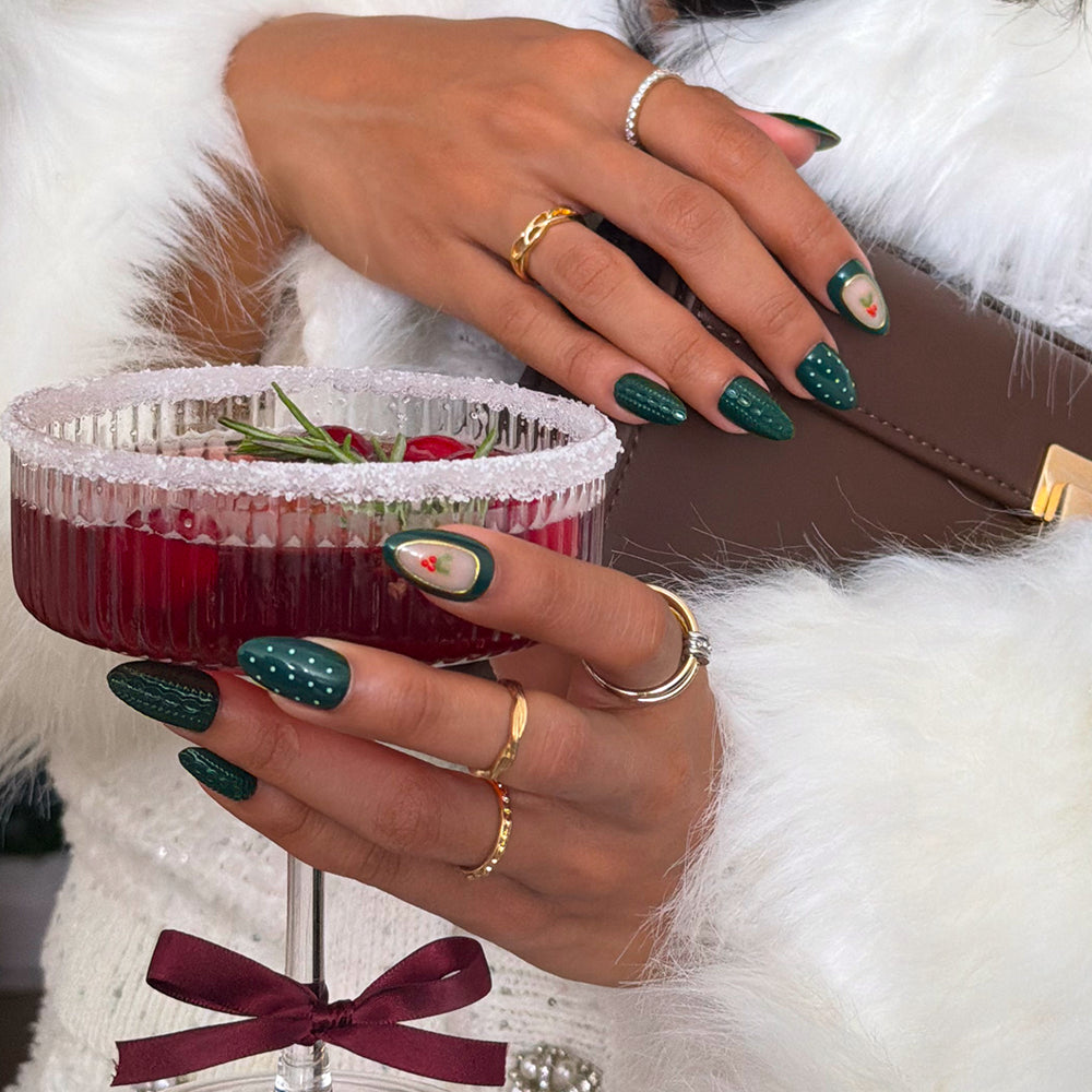 Hands wearing Table for Ten glamnetic press-on nails with short almond shapes, dark green polish, white polka dots, and red berry designs, holding a cocktail glass with a sugar-rimmed edge. - Product image