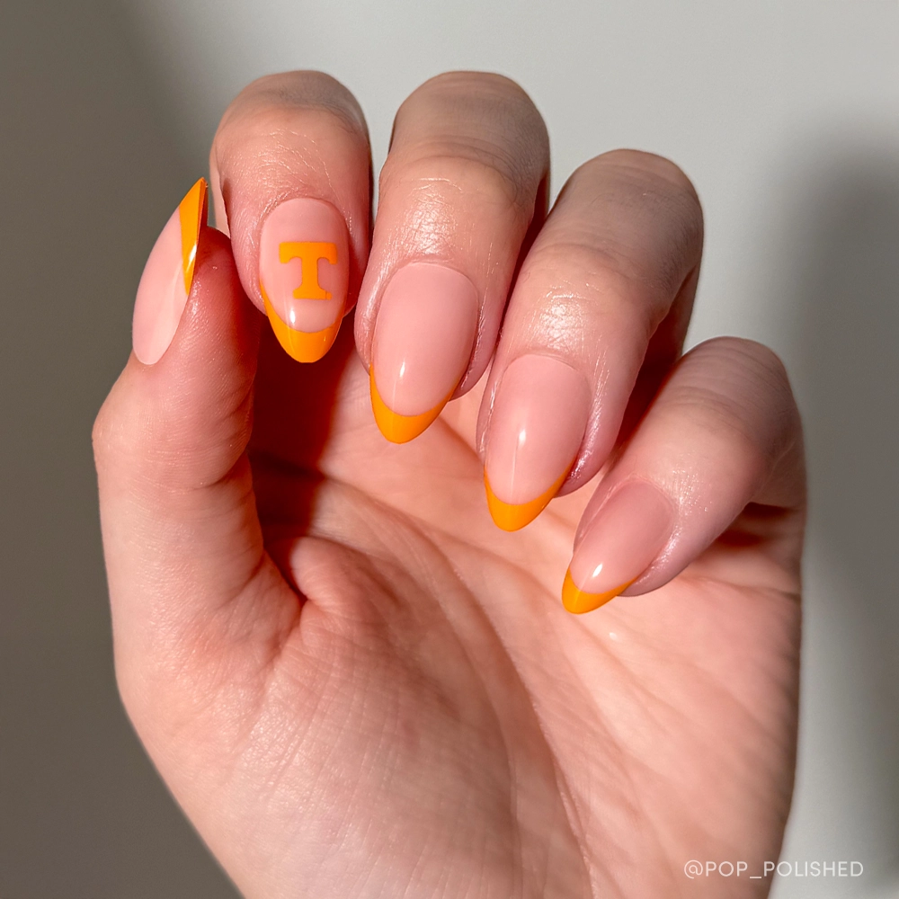A hand shows off the glamnetic Tennessee Volunteers Press-On Nail Set: almond-shaped with a nude base, bright orange French tips, and an orange T on the ring finger. Perfect Rocky Top nails in a well-lit, neutral background.