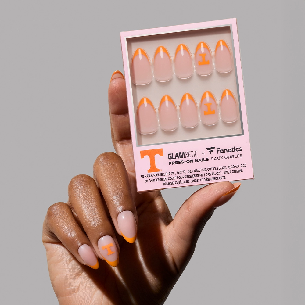 A hand with long, almond-shaped nails shows off the Tennessee Volunteers Press-On Nail Set by glamnetic, featuring orange French tips and a large orange T, while holding a box with matching Rocky Top nails inside.