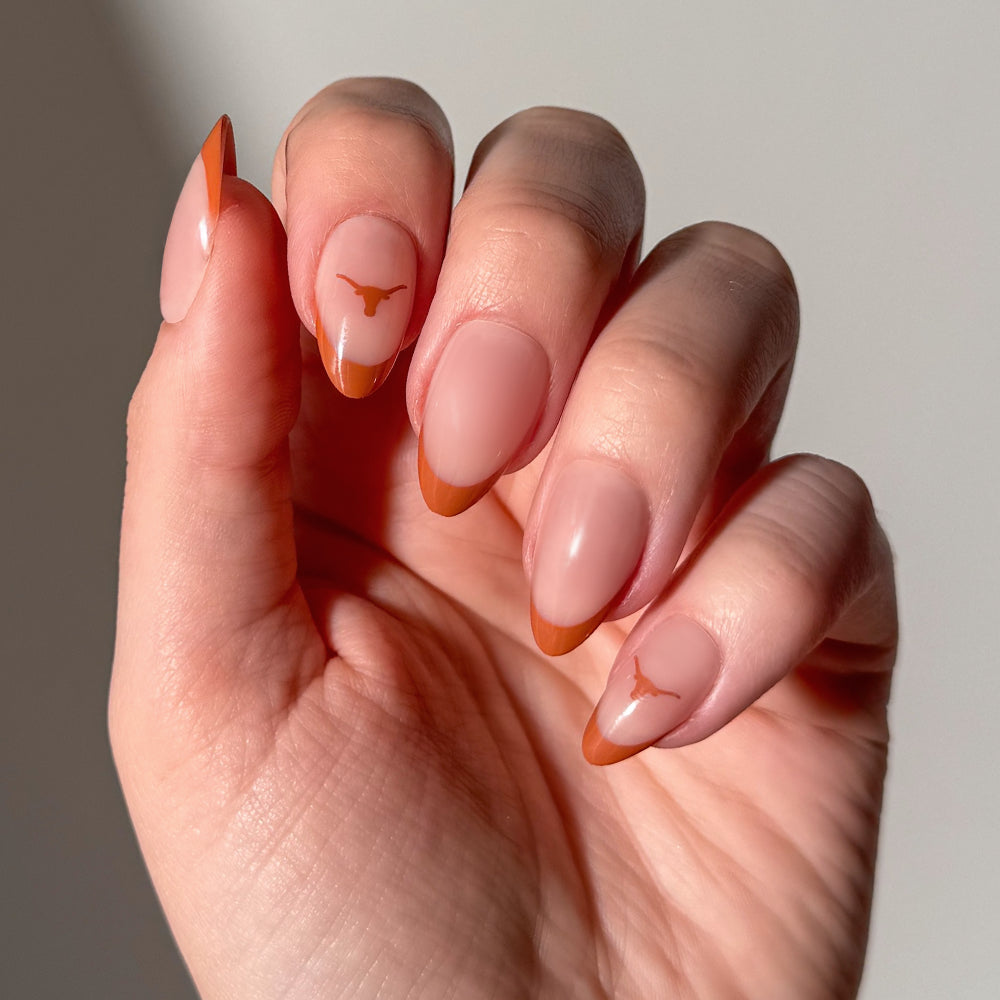 A hand wearing glamnetic Collegiate Press-On Nails, featuring almond-shaped nails with a light pink base, vibrant orange tips, and a small orange longhorn logo accenting two nails, displayed against a plain background. - Product image
