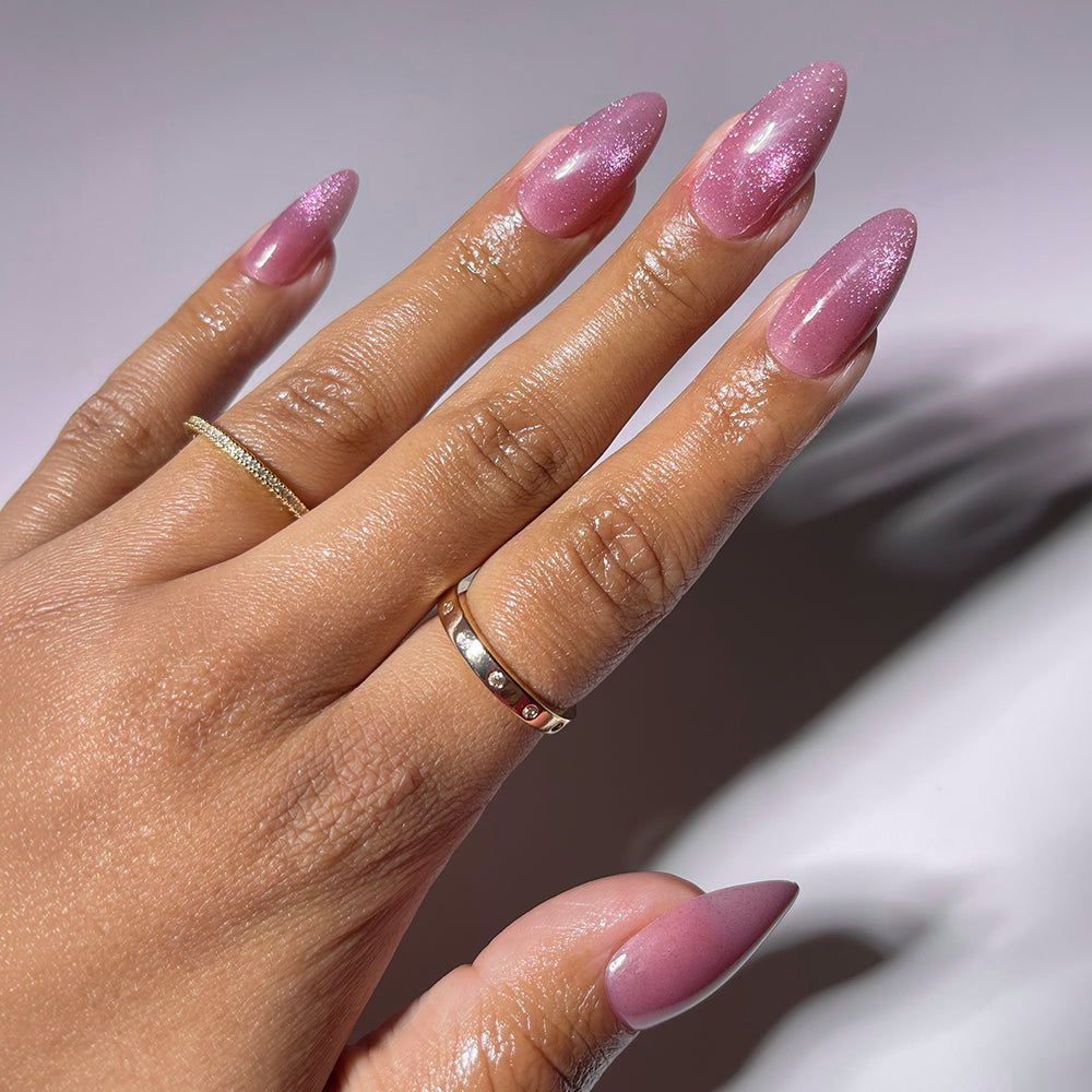 A moisturized hand wears glamnetic’s THE BEAUTY BLUEPRINT long, almond press-on nails in shimmery pink, paired with two gold rings—one plain, one with gems—resting against a light background.