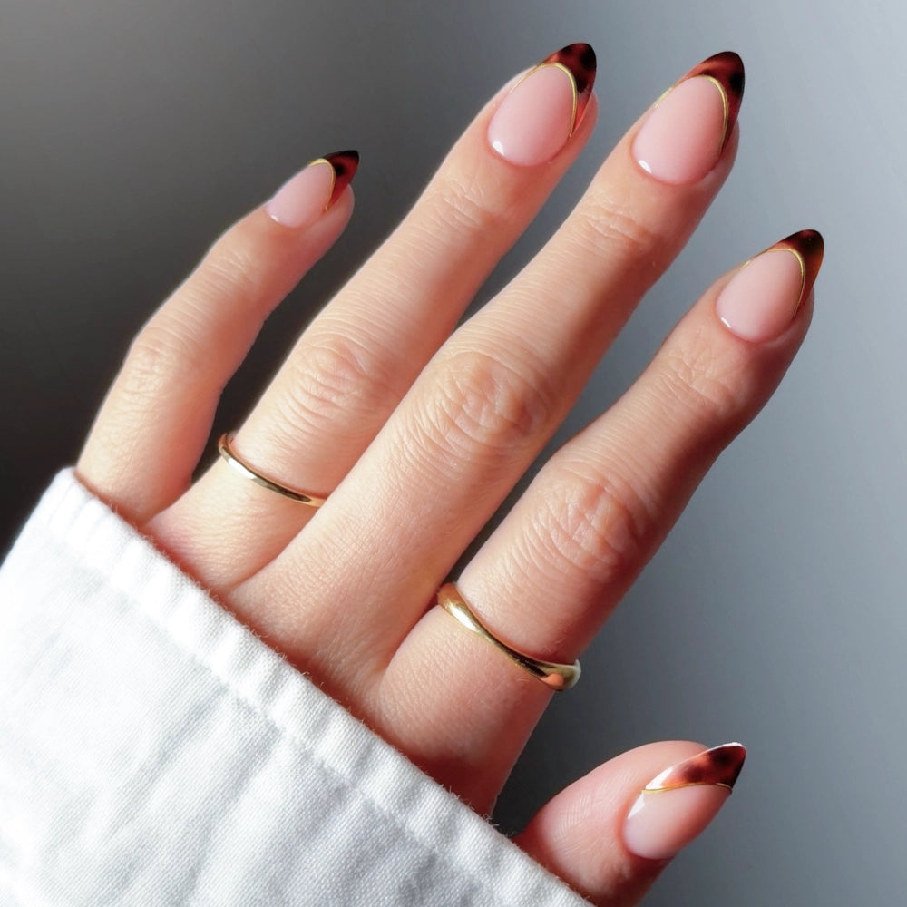 A hand with almond-shaped "The Original" by glamnetic nails, showcasing a pink base and tortoiseshell French tips, adorned with two gold rings and wearing a white long-sleeve garment. - Product image