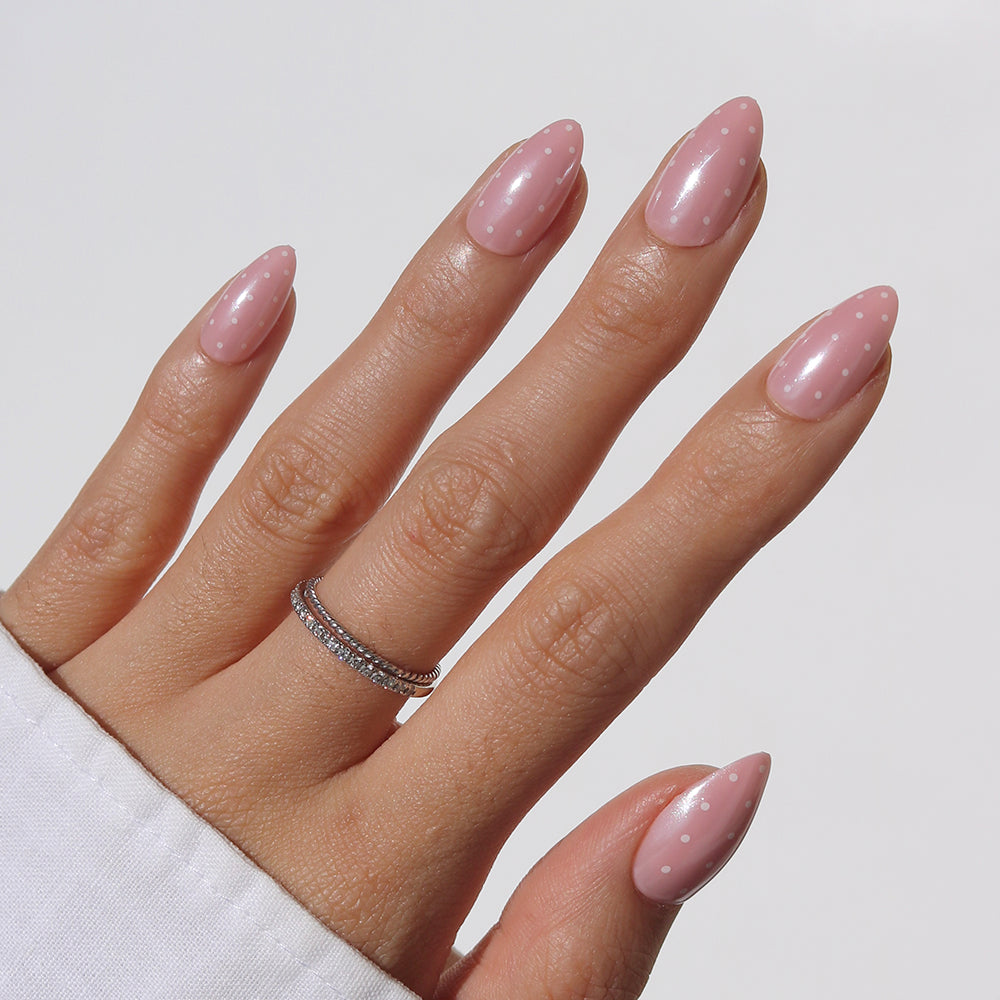 A hand shows off glamnetic’s Barely Spotted short almond nails in light pink with white polka dots, paired with a silver ring and white long sleeve top, set against a plain light background.