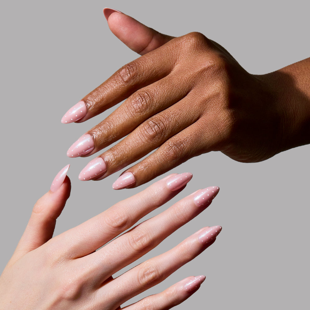 Two manicured hands—one dark-skinned, one light—display the glamnetic Barely Spotted nails. Each has short almond shapes in light pink with tiny white polka dots, set against a plain gray background for a chic, stylish look. - Product image