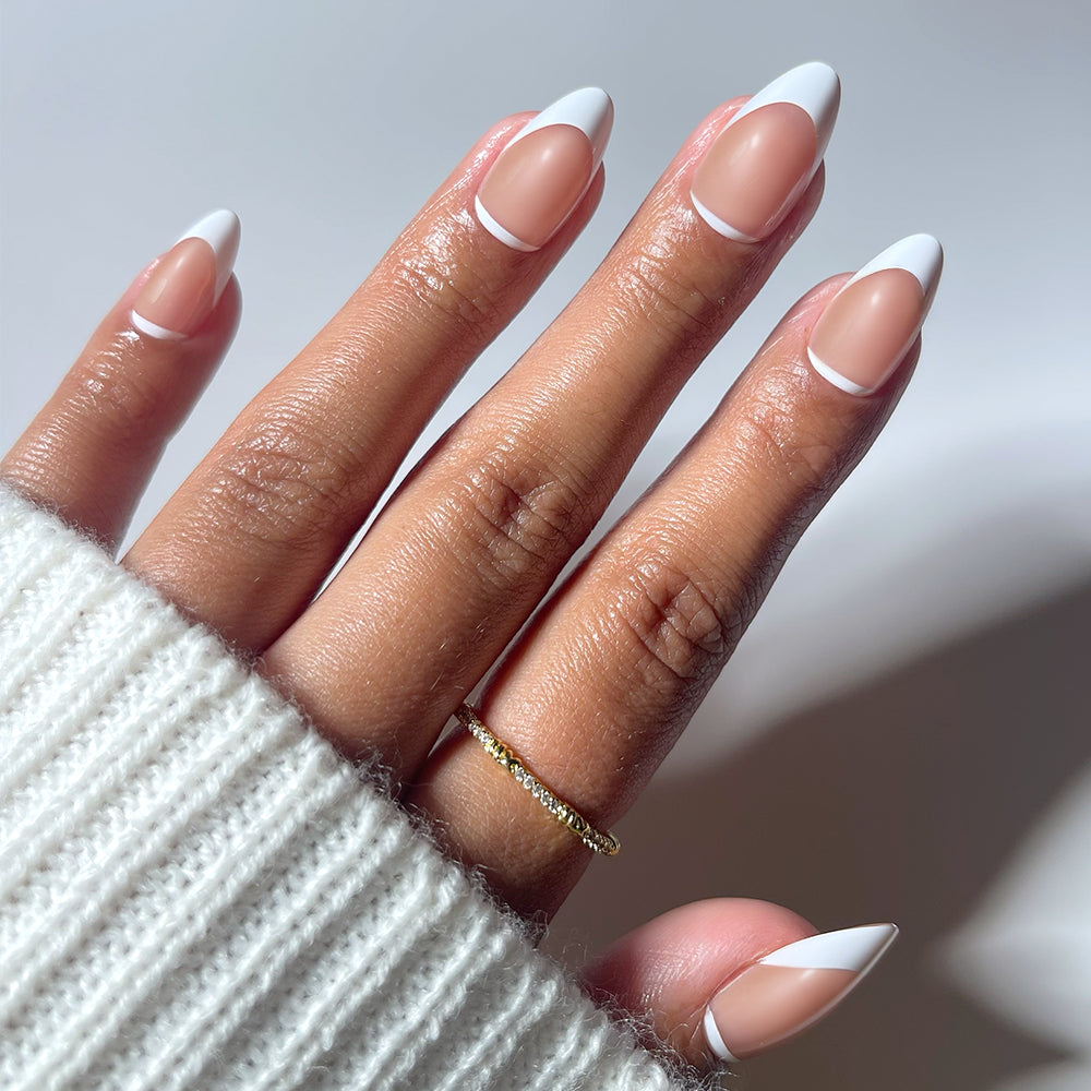 A hand with short almond nails wears glamnetic’s Class Act press-ons, showing a modern French manicure with white tips and a fine white line near the cuticles. A textured gold ring and a white knit sweater sleeve complete the chic look.