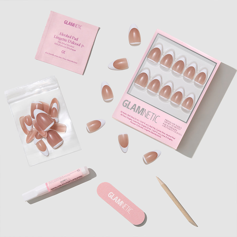The Class Act set by glamnetic includes pink-toned French tip short almond press-on nails, a nail file, wooden cuticle stick, nail glue, alcohol pad, and extra nails—all neatly arranged on a light gray background. - Product image