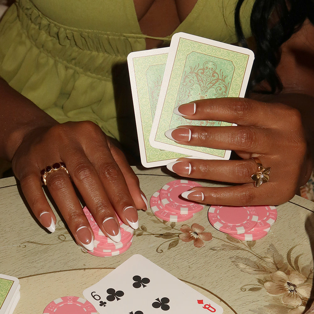 A person with short almond nails wearing glamnetic's Class Act holds playing cards, surrounded by pink poker chips and a six of clubs on the table. - Product image