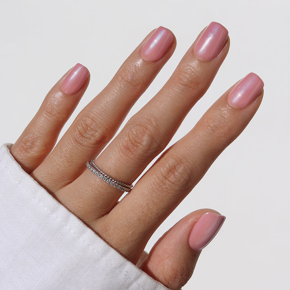 A hand with short squoval nails painted in glamnetic's Mauve On Up, a glossy mauve pink shade, wears a double-band silver ring with small stones. The nails are shown against a plain white background and the person is wearing a white long-sleeve top.