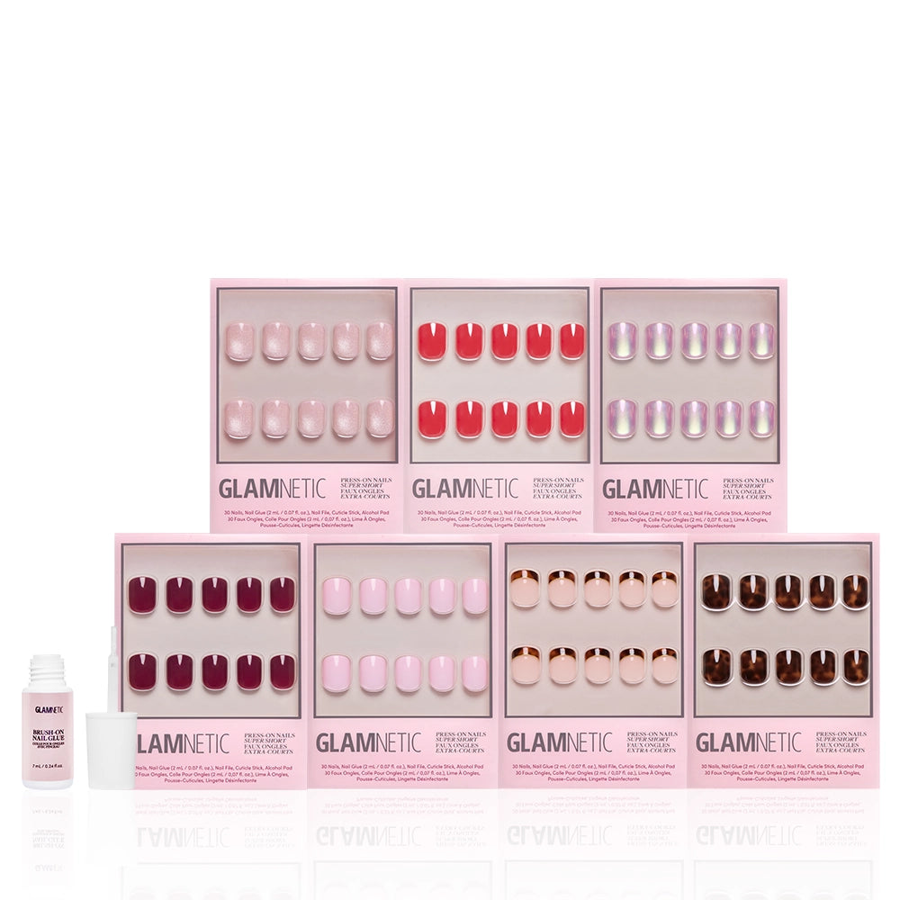 Seven boxes of glamnetic's The Super Short Edit glossy press-on nails, in pink, red, and neutral shades, are arranged in two rows with a small white nail glue bottle in front, displayed on a white background. - Product image