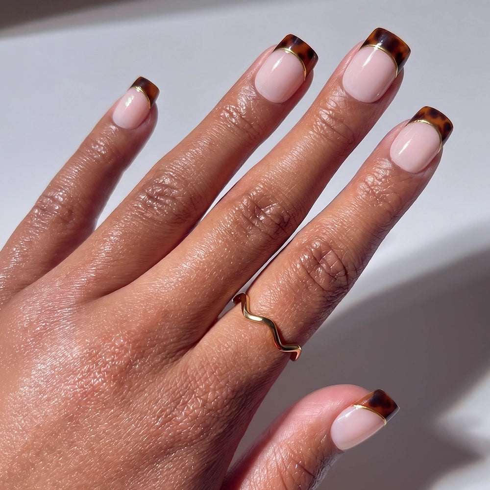 A hand wears glamnetic’s The Trendsetter: short squoval nails with a glossy nude base and dark brown tortoiseshell French tips, accented by a simple gold wavy ring on the middle finger.