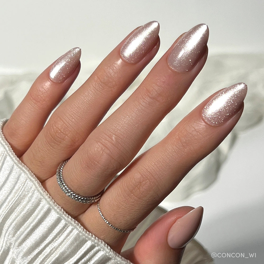 A hand with almond-shaped, shimmery pale pink The Vow nails by glamnetic, finished with a velvet-gloss sheen and paired with silver rings, rests on a white ribbed sleeve against a softly blurred background for an elegant look. - Product image
