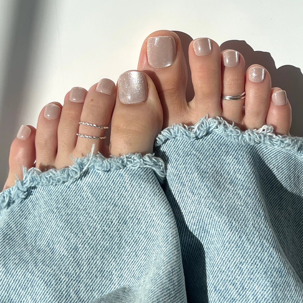 Close-up of feet with shimmering champagne silver Glamnetic press-on toenails, styled with silver rings and light-washed frayed denim jeans. - Product image
