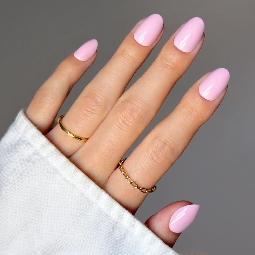 A hand with almond-shaped, glossy "Think Pink" nails by glamnetic wears two gold rings on the middle and ring fingers, set against a neutral background. The person is dressed in a white long-sleeve top.