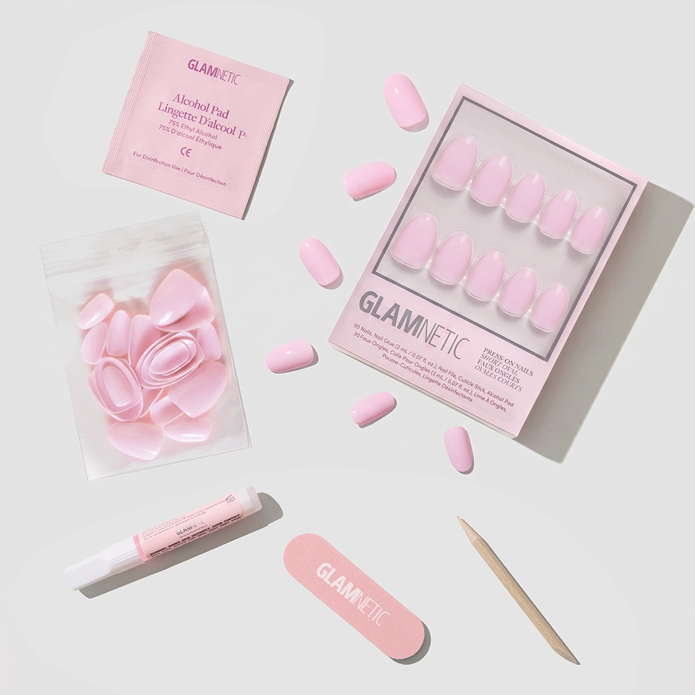 A flat lay of glamnetic's Think Pink press-on nail kit shows short oval baby pink nails, a nail file, wooden cuticle stick, alcohol pad, nail glue, and extra nails on a light background. - Product image