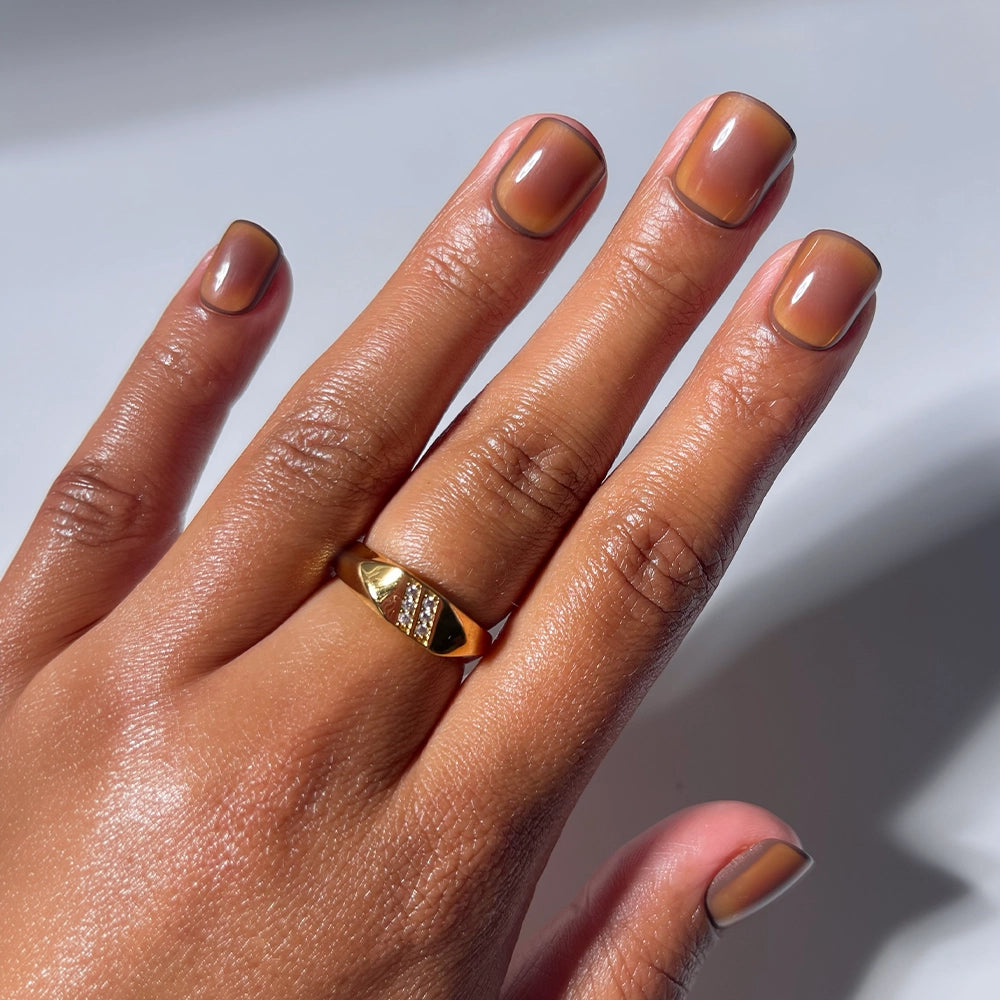 A hand with brown skin showcases the glamnetic Tiger Eye gold ring, featuring a geometric design. The super short nails are manicured with salon-quality finish—a natural base accented by a thin dark brown outline along the tips and sides—ideal for fans of minimalist nail art.