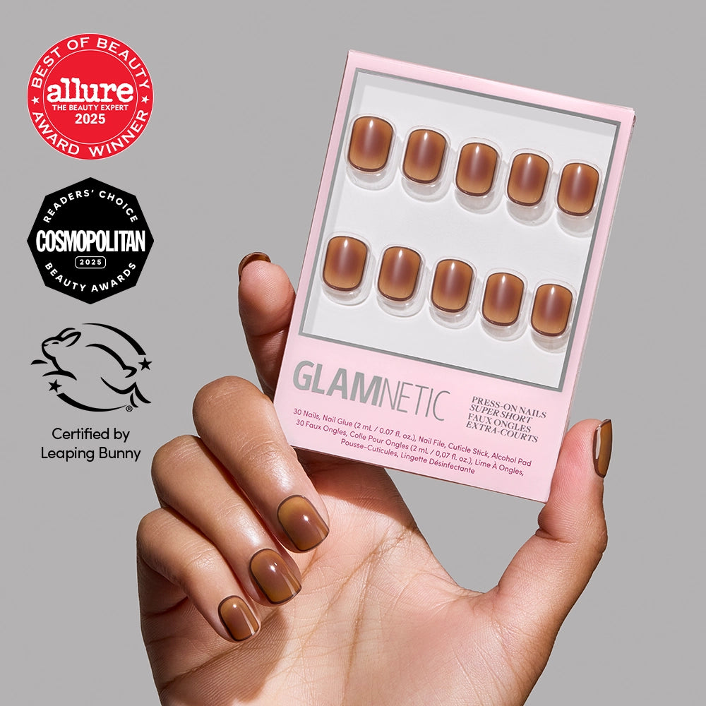 A hand with glossy brown Tiger Eye by glamnetic super short press-on nails holds a matching box. Allure, Cosmopolitan, and Leaping Bunny award badges appear on a light gray background.