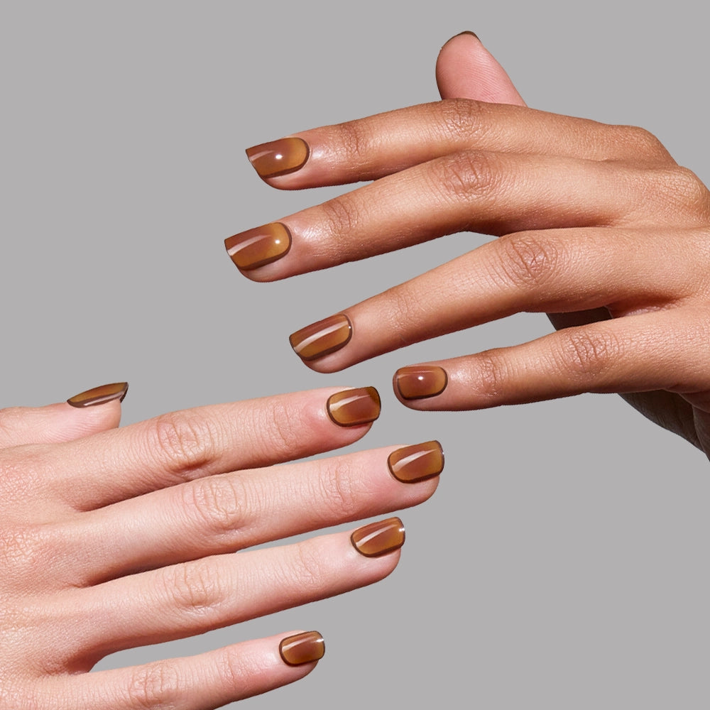 Two hands with super short glamnetic Tiger Eye press-on nails, neatly manicured and painted in a glossy brown shade, are posed against a plain gray background for salon-quality wear.
