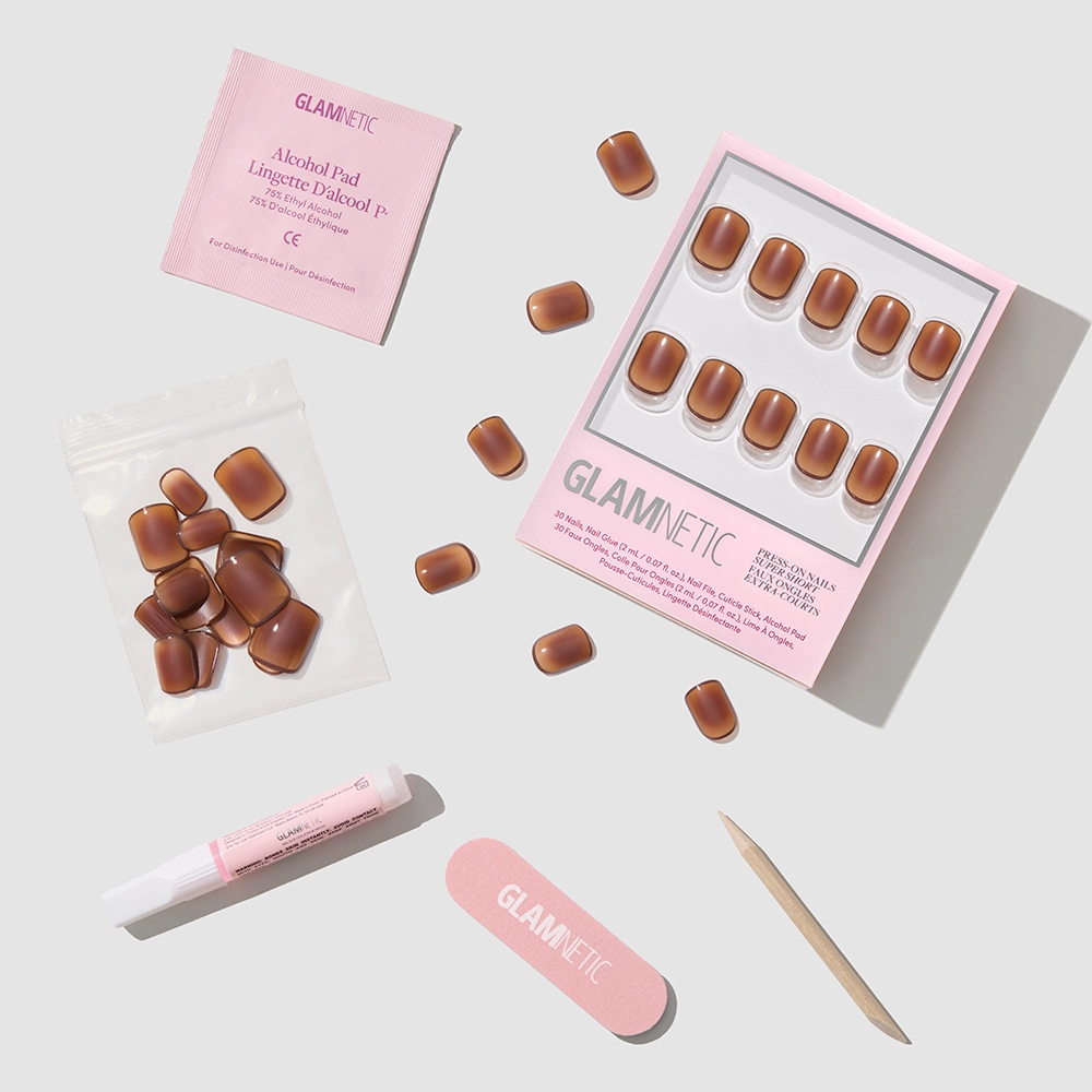 The Glamnetic Tiger Eye kit includes super short press-on nails, nail glue, an alcohol pad, a pink nail file, a cuticle stick, and extra nails, all arranged neatly on a light gray surface for salon-quality results. - Product image
