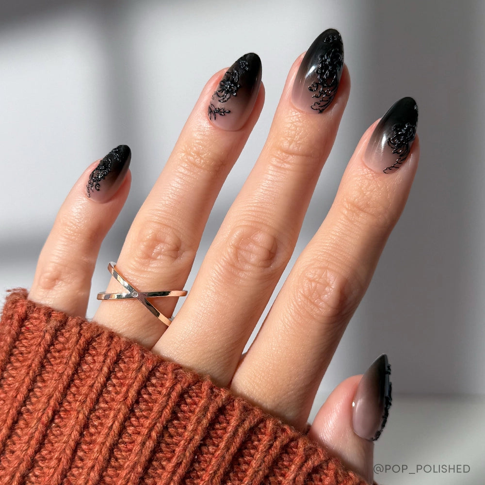 A hand with black almond nails featuring a black ombre and lace accent, adorned with a crisscross gold ring and an orange-brown ribbed sweater—showcasing the stunning ‘Til Death set by glamnetic.