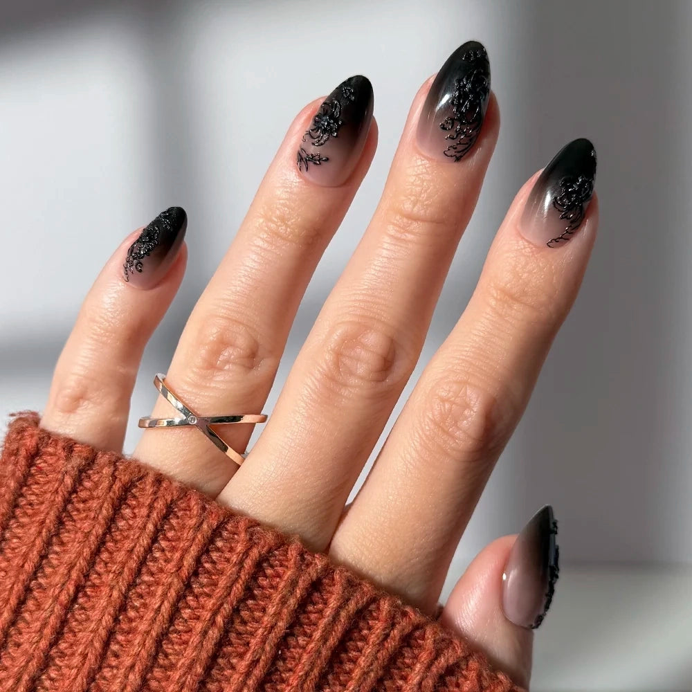 A hand with glossy ‘Til Death nails by glamnetic in a long almond shape, featuring black ombre tips and floral details, wears a gold crisscross ring and a rust-colored knit sweater.