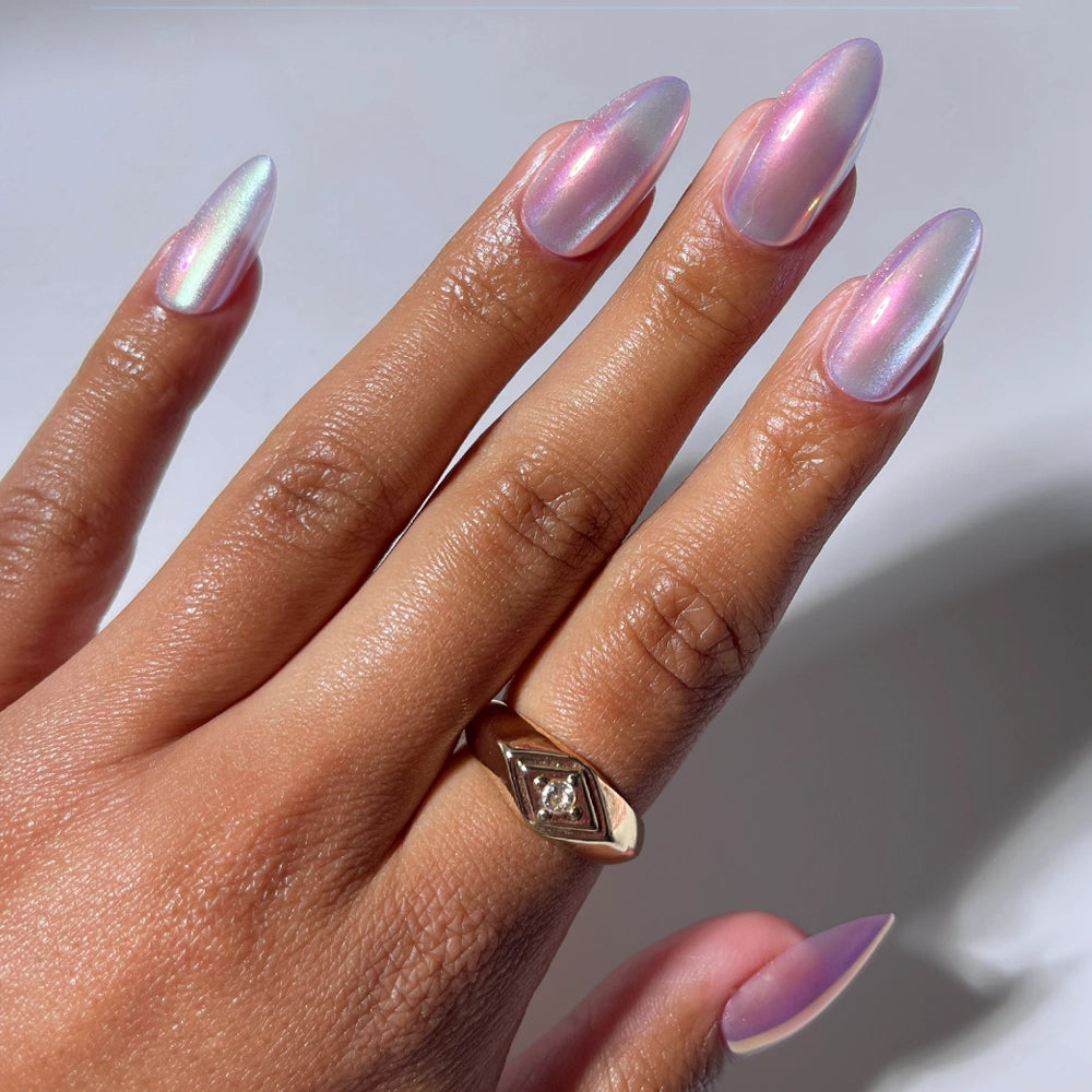 A hand with long, almond-shaped glamnetic Timeless Oyster press-on nails in pearlescent, iridescent light pink polish, wearing a gold ring with a diamond on the ring finger.