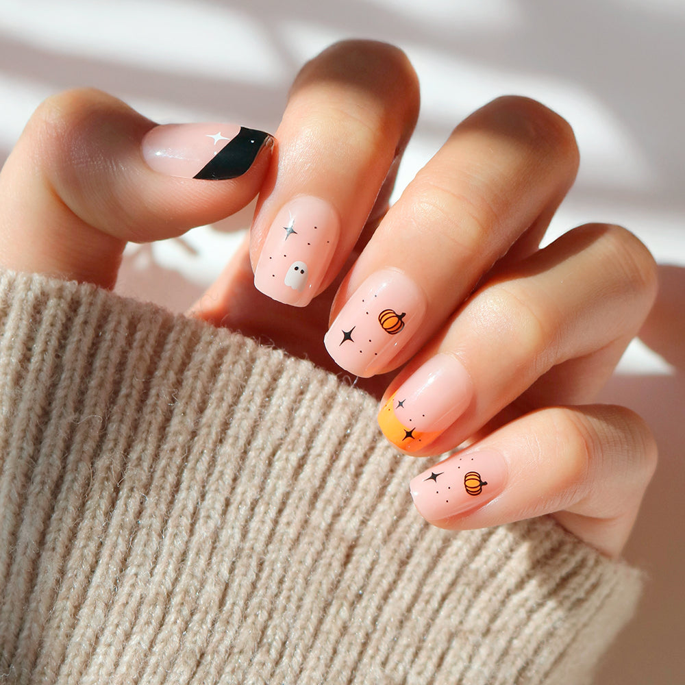A hand with long, manicured nails featuring glamnetic’s Tiny Pumpkin Halloween nail art—complete with playful stars, planets, and abstract shapes—rests against a soft beige sweater background.