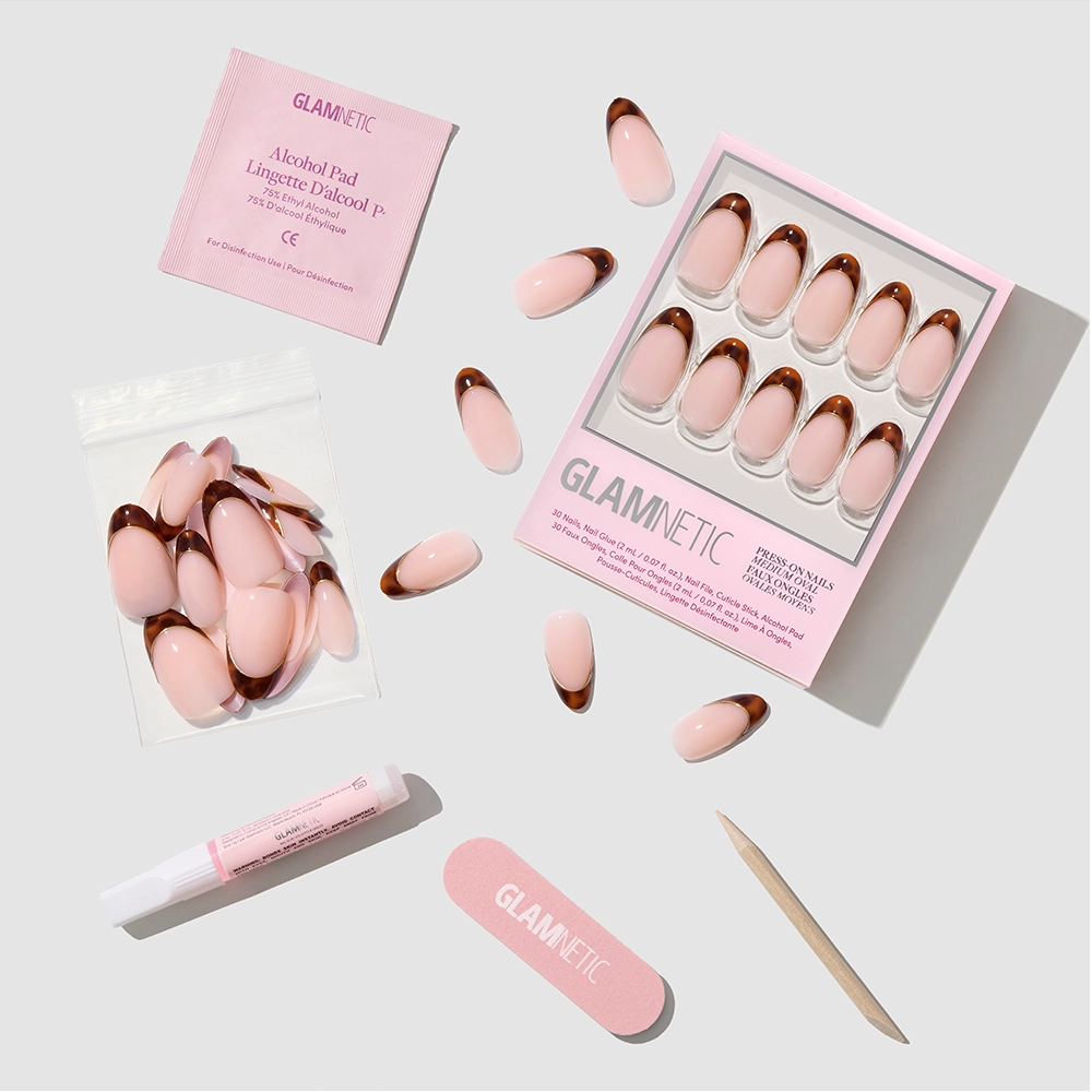 A flat lay of the glamnetic "Too Much" press-on nail kit shows oval pink and brown French tip nails, alcohol pad, nail glue, file, cuticle stick, and extra nails arranged on a white background. - Product image